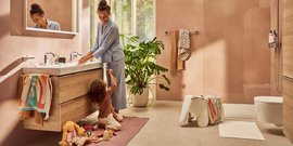 Eine Mutter lächelt, während sie am Waschbecken steht, während ihr Kind auf dem Boden mit Spielzeug spielt.