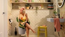 Eine Frau mit einer Gesichtsmaske sitzt entspannt auf der Toilette und schaut auf ihr Handy, während sie ein Magazin hält.